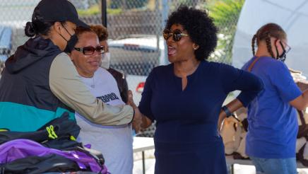 Assemblymember McKinnor shakes a volunteers hand while they stand next to a pile of backpacks to give away