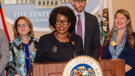 Asm. McKinnor at podium, speaking, with fellow legislators standing in support