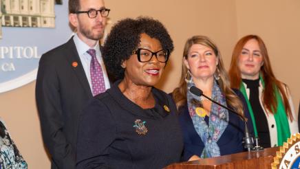 Asm. McKinnor at podium, speaking, with fellow legislators and guest speakers standing in support