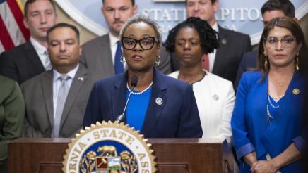 Asm. McKinnor at podium, speaking, with fellow legislators standing in support
