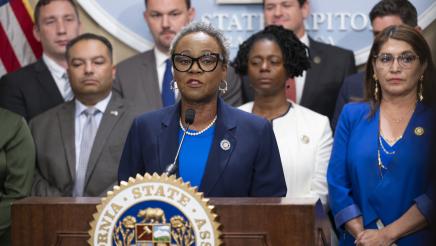 Asm. McKinnor at podium, speaking, with fellow legislators standing in support