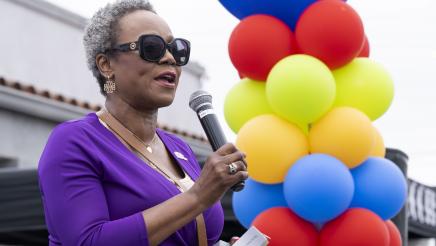 Asm. McKinnor holding microphone, speaking, with column of ballons in background