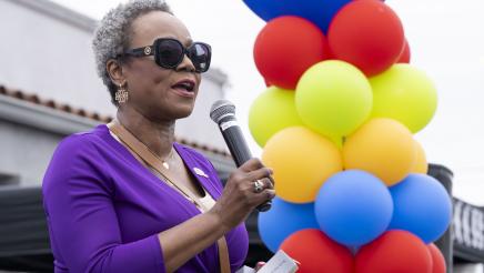 Asm. McKinnor holding microphone, speaking, with column of ballons in background